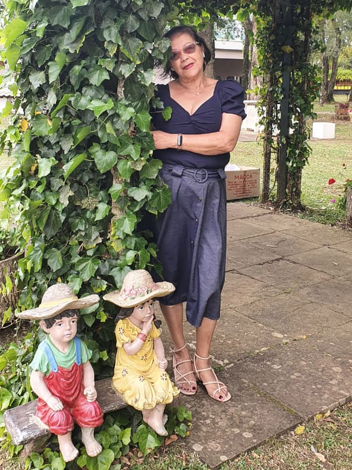 Lydia sorrindo, de braços cruzados, ao lado de um arco de hera no jardim