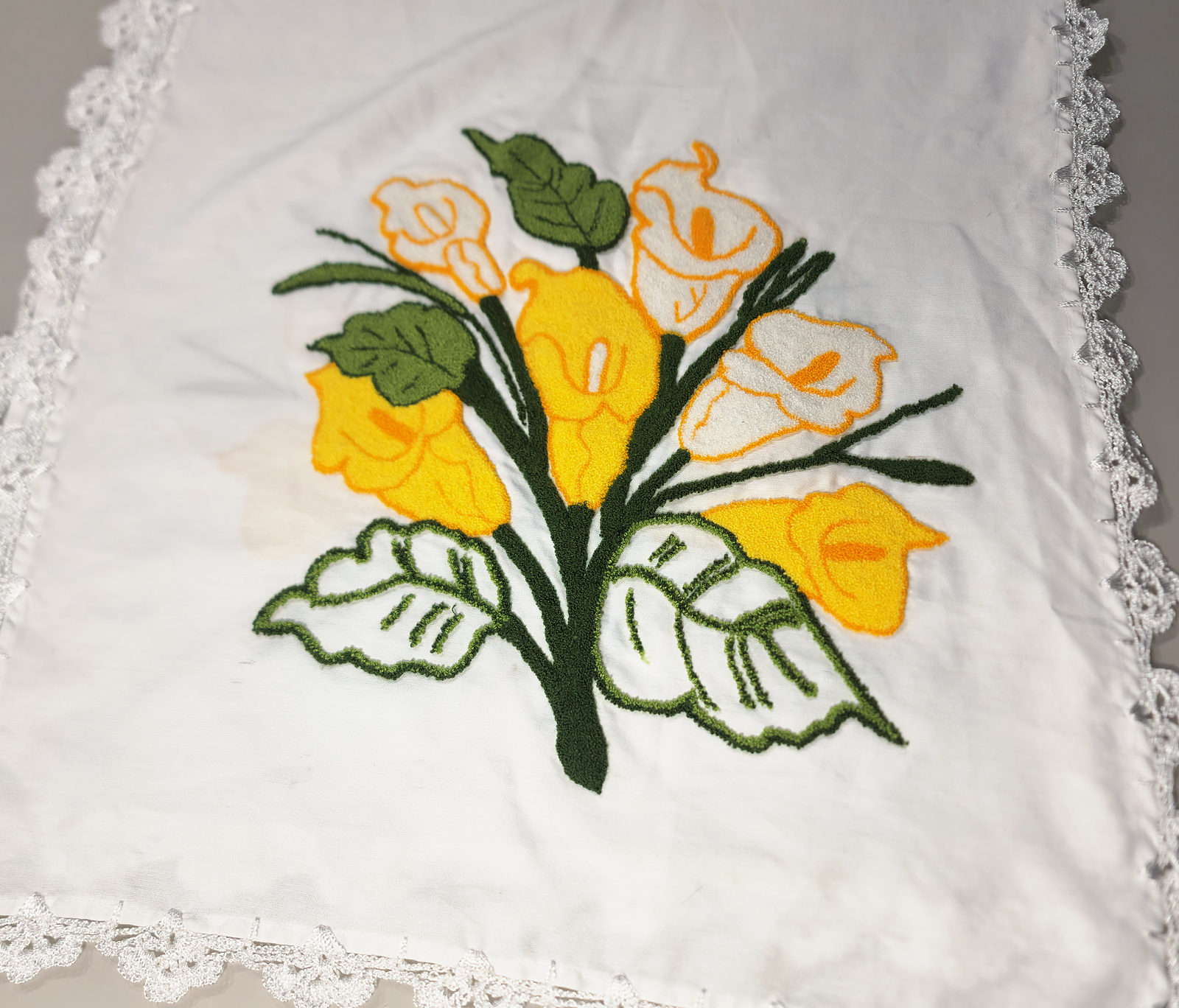 Pano branco bordado com um buquê de copos-de-leite amarelos e brancos e folhas verdes, com barrado de crochê branco