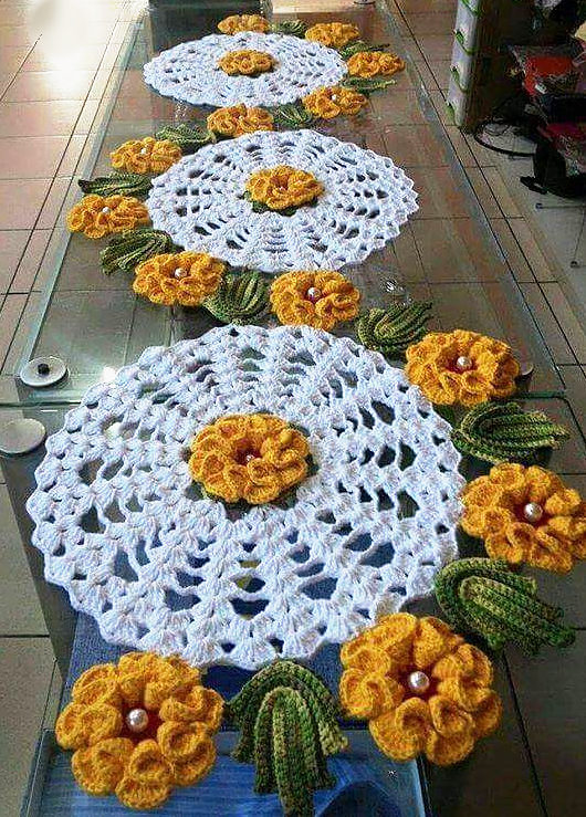 Centros de mesa redondos de crochê branco com flores amarelas e folhas, enfileirados sobre uma mesa de vidro