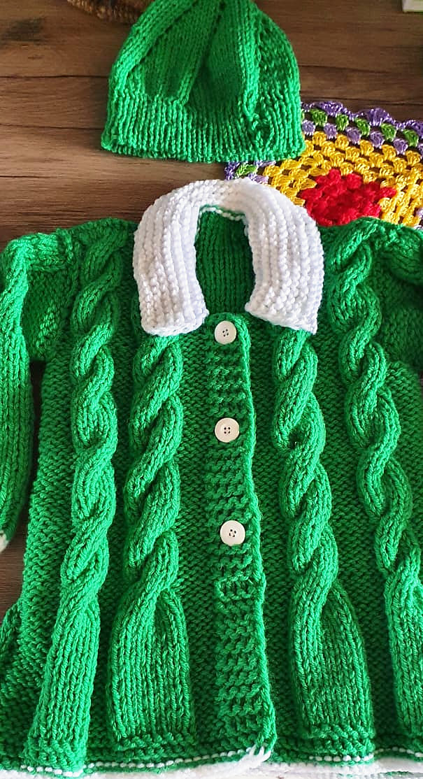 Casaquinho infantil verde de tricô com tranças, gola branca e botões, com a touca verde combinando